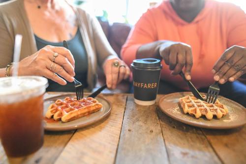 Two people sit at a wooden table, enjoying waffles and coffee, each using a fork to cut their waffles.