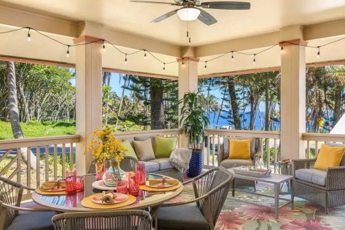 A cozy outdoor seating area with a dining table, surrounded by greenery and ocean views, decorated with colorful cushions.
