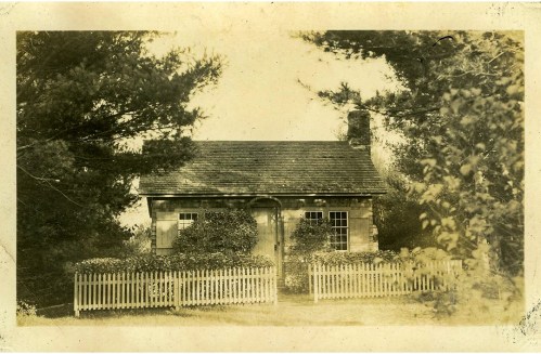 A vintage photo of a small house surrounded by trees and a white picket fence.