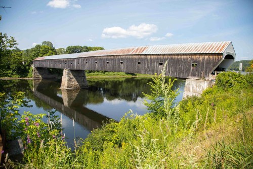 A weathered covered bridge spans a calm river, surrounded by lush greenery and a clear blue sky.