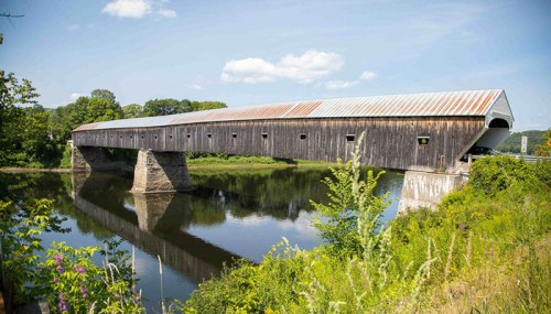 A long, wooden covered bridge spans a calm river, surrounded by greenery and blue skies.