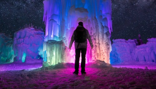 A person stands in front of colorful ice formations illuminated under a starry night sky.