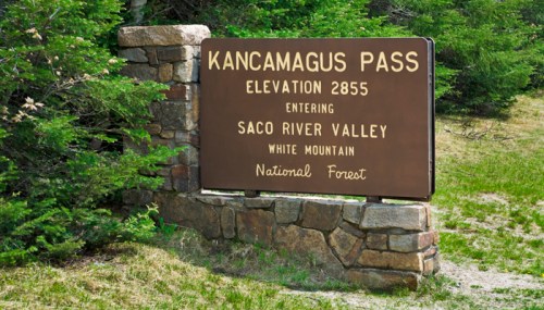 Sign for Kancamagus Pass, elevation 2855, entering Saco River Valley, White Mountain National Forest, surrounded by greenery.