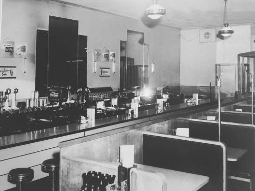 Black and white photo of a vintage diner interior with a long counter and booths, featuring retro decor and lighting.