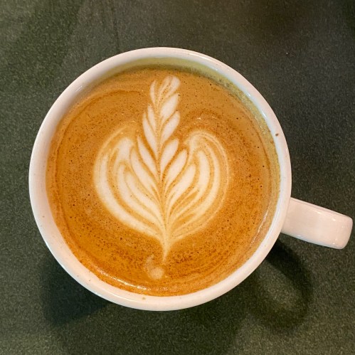 A close-up of a latte in a white cup, featuring intricate leaf-shaped foam art on top.