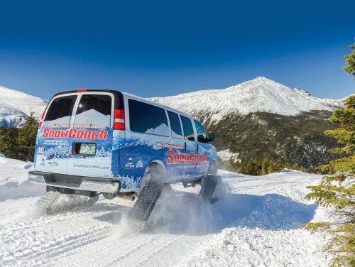 A snow coach van drives through a snowy landscape with mountains in the background under a clear blue sky.
