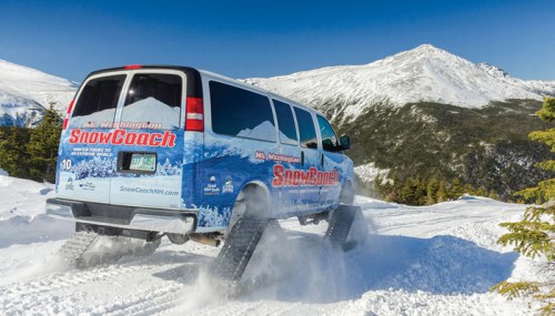 A snow coach van drives through a snowy landscape with mountains in the background under a clear blue sky.