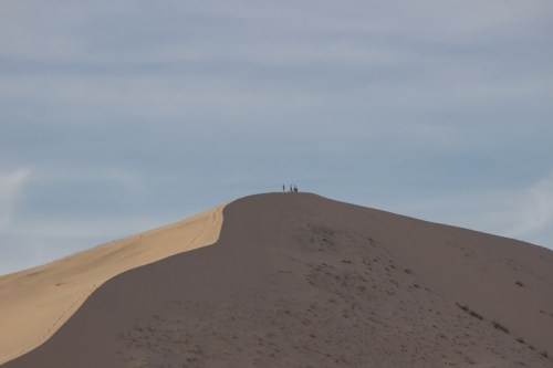A sandy dune with a few people standing at the top against a blue sky.