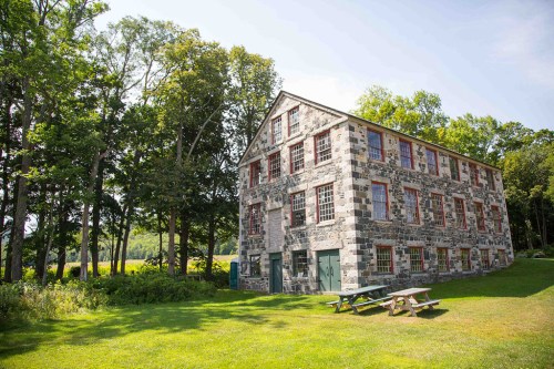 A large stone building with red windows, surrounded by trees and a grassy area with picnic tables.