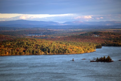 A scenic view of a lake surrounded by colorful autumn foliage and distant mountains under a cloudy sky.