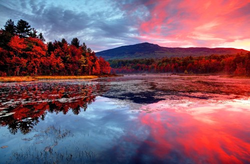 Vibrant sunset over a calm lake, reflecting red and orange hues with autumn trees and a mountain in the background.