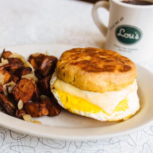 A plate with a biscuit sandwich containing an egg and cheese, served with crispy potato cubes and a coffee cup in the background.