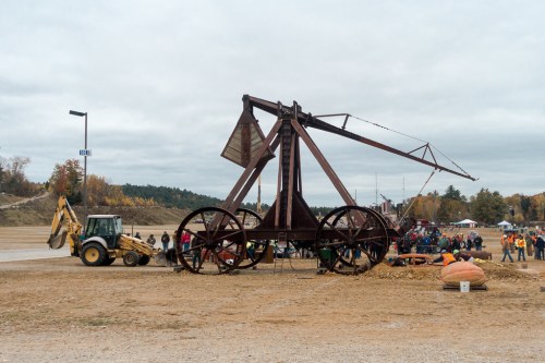 A large, vintage crane stands in a field with a crowd nearby, while a tractor operates beside it under a cloudy sky.