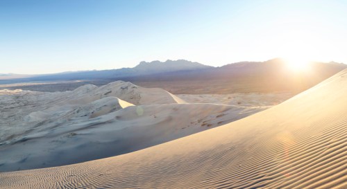 A serene desert landscape at sunrise, with rolling sand dunes and distant mountains under a clear blue sky.
