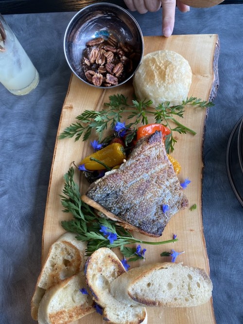 A wooden platter with grilled fish, roasted vegetables, a biscuit, and pecans, garnished with herbs and edible flowers.