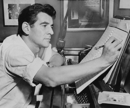 A composer writes music at a piano, focused on a sheet of music with notes and a pencil in hand.