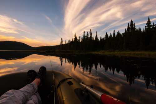 A person relaxes in a kayak on a calm lake, surrounded by trees and reflecting colorful clouds at sunset.