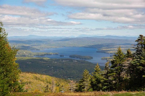 A panoramic view of a lake surrounded by lush forests and distant mountains under a partly cloudy sky.