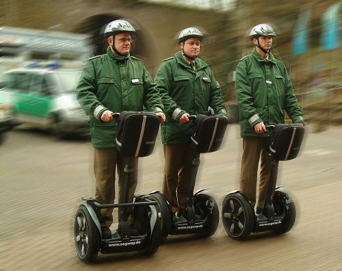 Three men in green uniforms and helmets riding Segways on a paved surface.
