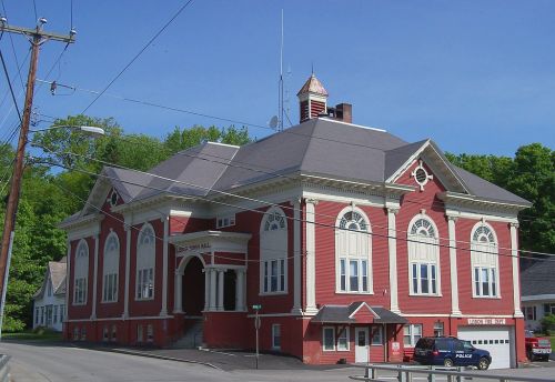 Historic red building with large windows and a clock tower, surrounded by trees and power lines.