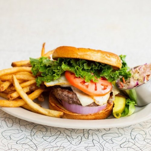 A juicy burger with lettuce, tomato, and onion, served with crispy fries and a side of coleslaw on a patterned table.