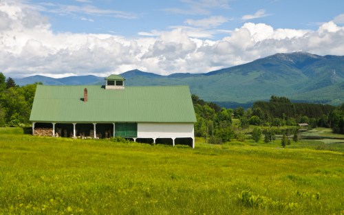 A white barn with a green roof sits in a lush green field, surrounded by mountains and a blue sky with clouds.