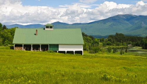 A green-roofed barn sits in a lush field, surrounded by mountains and a blue sky with fluffy clouds.