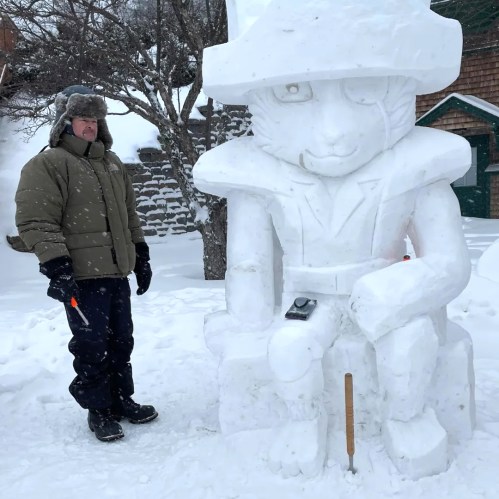 A person in a winter coat stands next to a large snow sculpture of a seated figure wearing a hat. Snowy landscape in background.