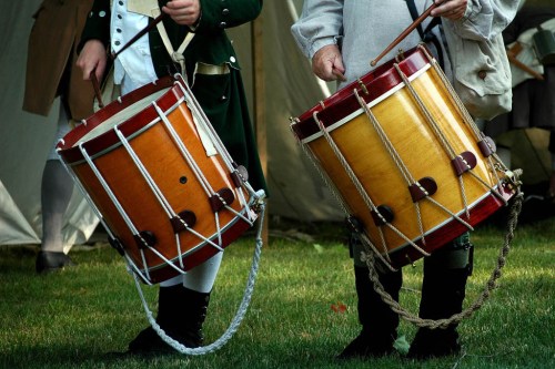 Two drummers hold traditional wooden drums with red and yellow finishes, set against a grassy background.