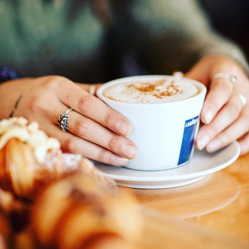 A person’s hands holding a cup of coffee with a sprinkle of cinnamon, resting on a saucer, with pastries in the foreground.