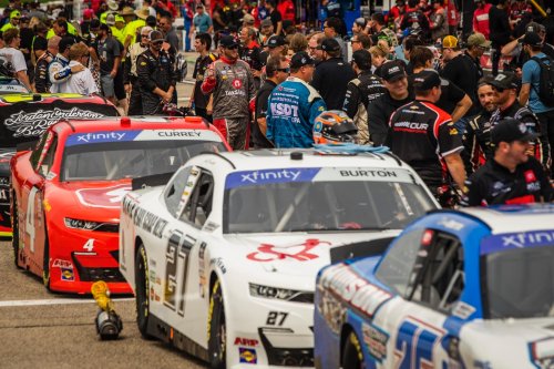 A busy NASCAR pit area with cars and crew members preparing for a race, surrounded by fans and team personnel.