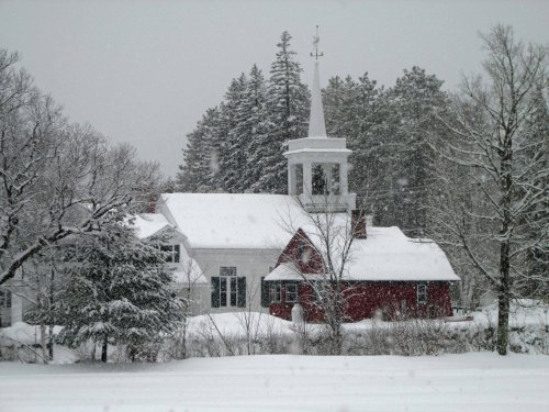 A red church with a white steeple surrounded by snow-covered trees in a winter landscape. Snow is falling gently.
