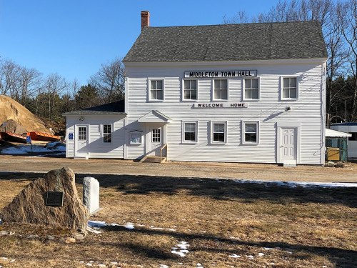 A white two-story building labeled "Middleton Town Hall" with a welcome sign, surrounded by grass and a stone monument.