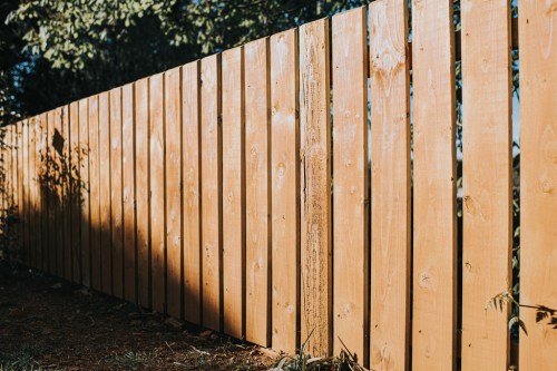 A wooden fence made of vertical planks, casting shadows on the ground in a sunny outdoor setting.