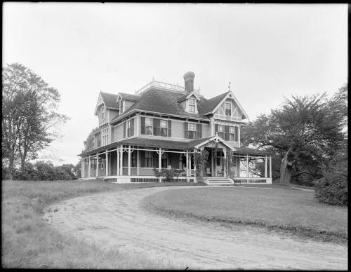 A large, historic house with a wraparound porch, surrounded by trees and a grassy area, captured in black and white.