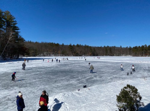 A frozen lake with people ice skating and enjoying winter activities under a clear blue sky. Snowy landscape surrounds the scene.