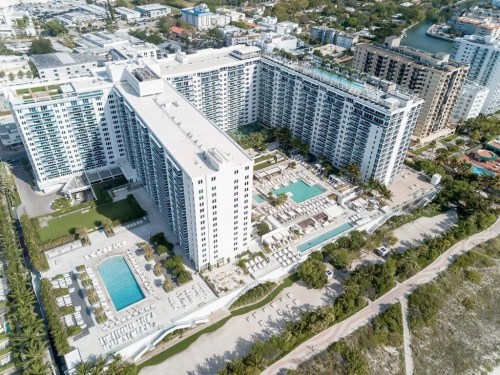 Aerial view of a large hotel complex with pools, palm trees, and surrounding buildings near a waterfront.