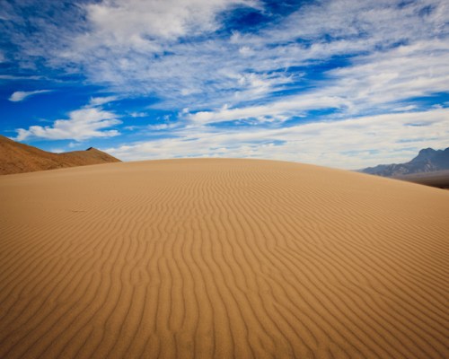 A vast desert landscape with rippled sand dunes under a blue sky with scattered clouds.