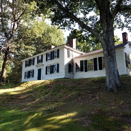 A white two-story house with black shutters, surrounded by trees on a grassy hillside.