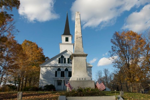 A white church with a tall steeple and an obelisk monument in front, surrounded by autumn trees and a blue sky.