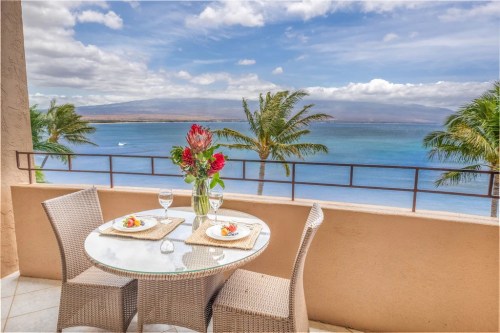 A scenic balcony with a glass table set for two, overlooking a blue ocean and palm trees under a cloudy sky.