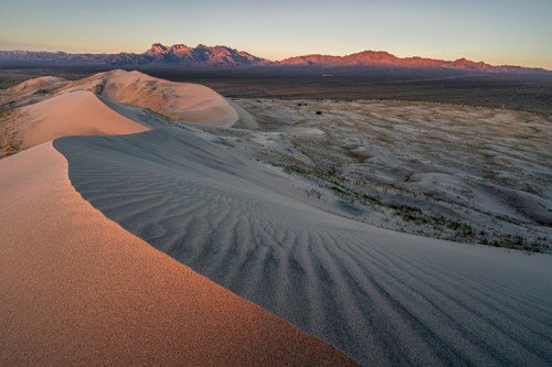 A serene desert landscape with rolling sand dunes and distant mountains under a soft sunset glow.