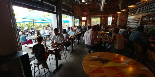 A bustling restaurant interior with diners at tables, large windows, and outdoor seating under blue umbrellas.