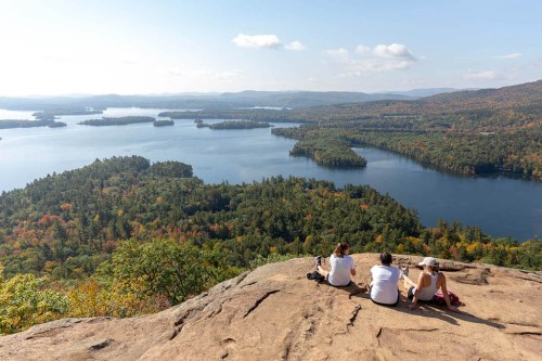 Three people sit on a rocky ledge, overlooking a scenic lake surrounded by colorful autumn foliage.
