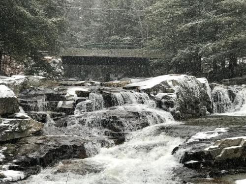 A snowy landscape featuring a flowing river over rocks, with a bridge visible in the background among the trees.