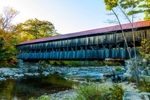 A wooden covered bridge spans a river, surrounded by autumn foliage and rocky banks under a clear blue sky.