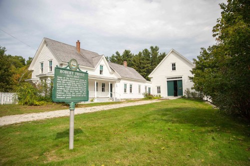 A historic house with a green sign marking it as Robert Frost's home, surrounded by trees and a grassy area.