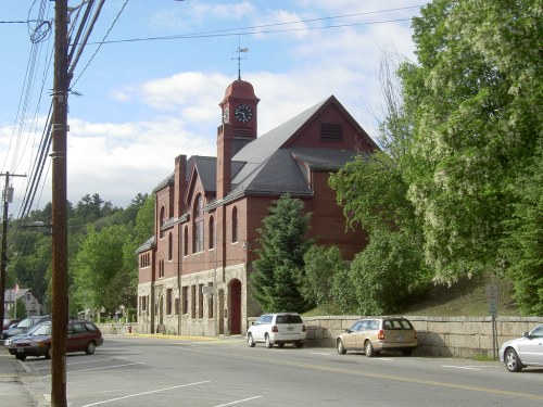 Historic red brick building with a clock tower, surrounded by trees and parked cars on a sunny day.