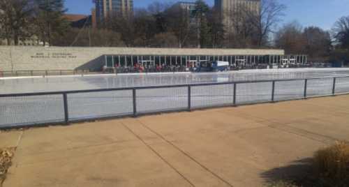 Outdoor ice skating rink with a building in the background, surrounded by trees and a clear blue sky.