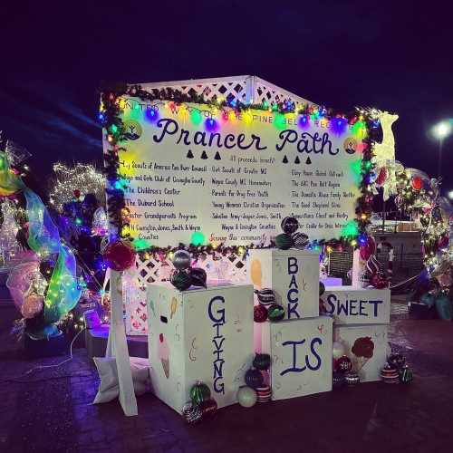 A festive display with colorful lights and decorations, featuring a sign that reads "Giving Back is Sweet" on Prancer Path.